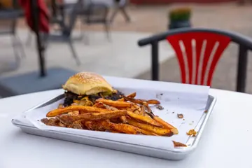 Una hamburguesa smash con queso derretido y un montón de papas en una charola con papel, sobre una mesa blanca del patio.