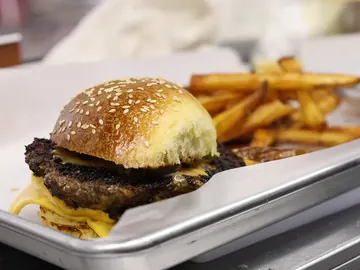 Una hamburguesa smash doble con queso americano derretido en pan con ajonjolí, con papas al lado, en una charola de acero con papel.