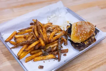 Un montón de papas sazonadas en una charola con papel, con una hamburguesa smash atrás.