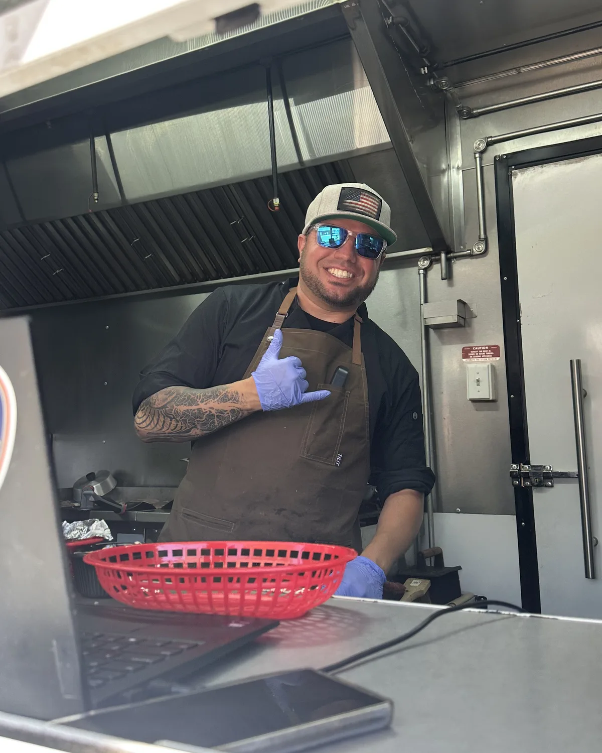 Joe Sutliff en la ventana del tr&aacute;iler de Chef Joe, sonriendo con gorra y mandil y haciendo el saludo shaka.