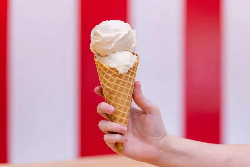 Una mano sostiene un cono de waffle con dos bolas de helado de vainilla de Mimi&rsquo;s, frente a rayas rojas y blancas.